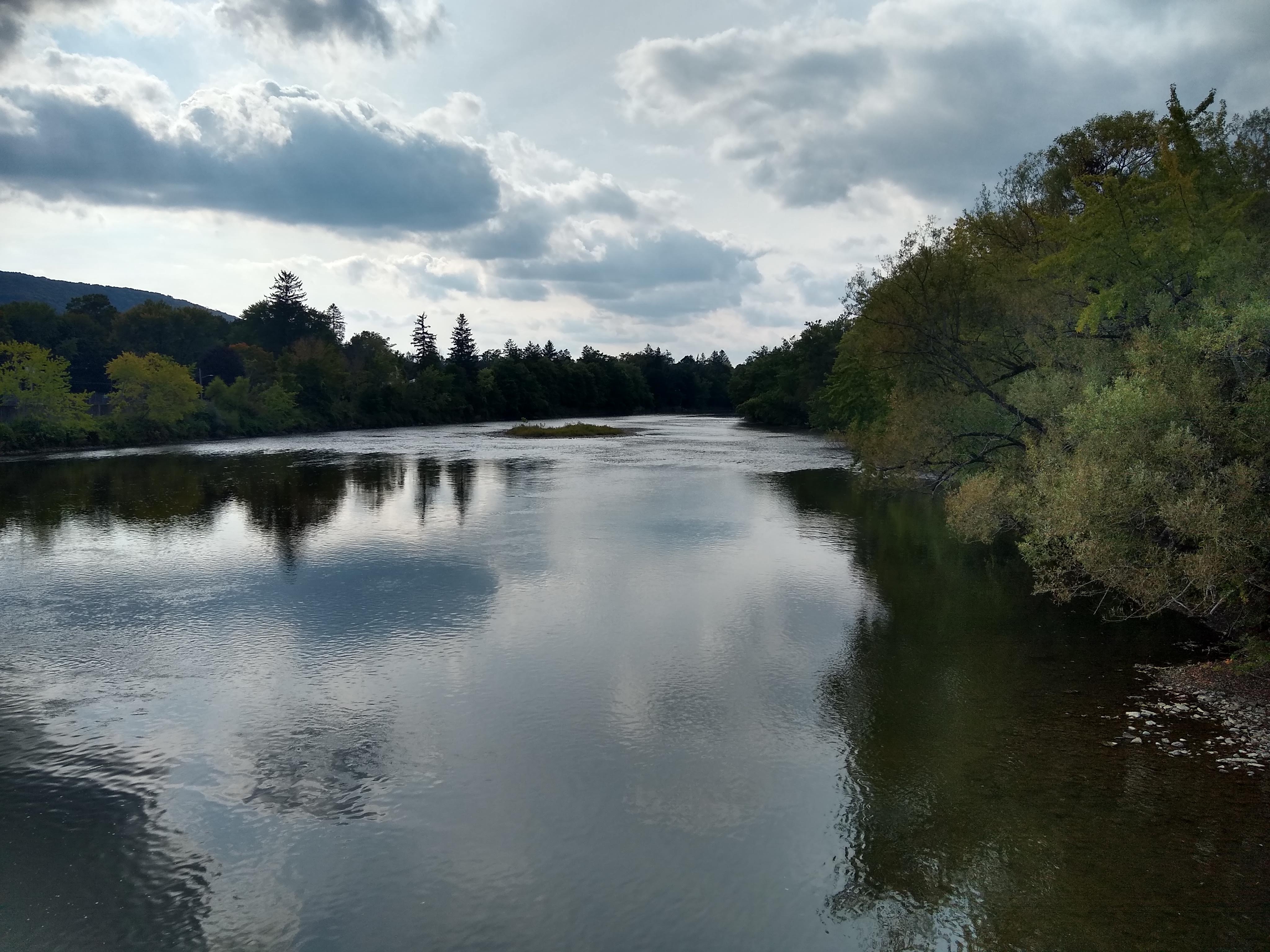 A view of Ohi:yo’, the Allegany River, in Salamanca, NY, with trees on both banks, with the sun and clouds in the sky reflecting in the water
